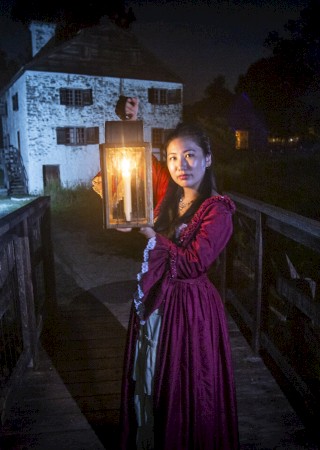 A person in period clothing holds a lantern on a bridge at night, with an old stone building in the background under eerie lighting.