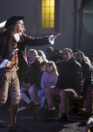 A person in historical attire performs in front of a seated audience, with dim lighting creating a dramatic ambiance.