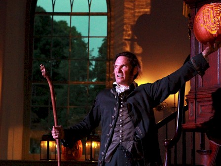 A person in historical attire holds a carved pumpkin in one hand and a staff in the other, standing dramatically in dim light.