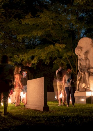 A group of people gathers at night around a statue in a park, illuminated by lights, surrounded by trees.