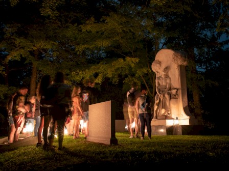 A group of people gathers at night around a statue in a park, illuminated by lights, surrounded by trees.