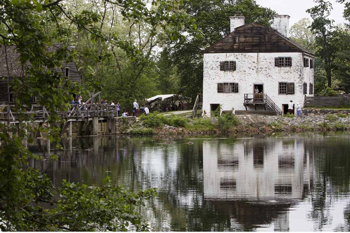 The image shows a historic white building reflecting in a pond, surrounded by trees. A bridge and people are visible on the left side.