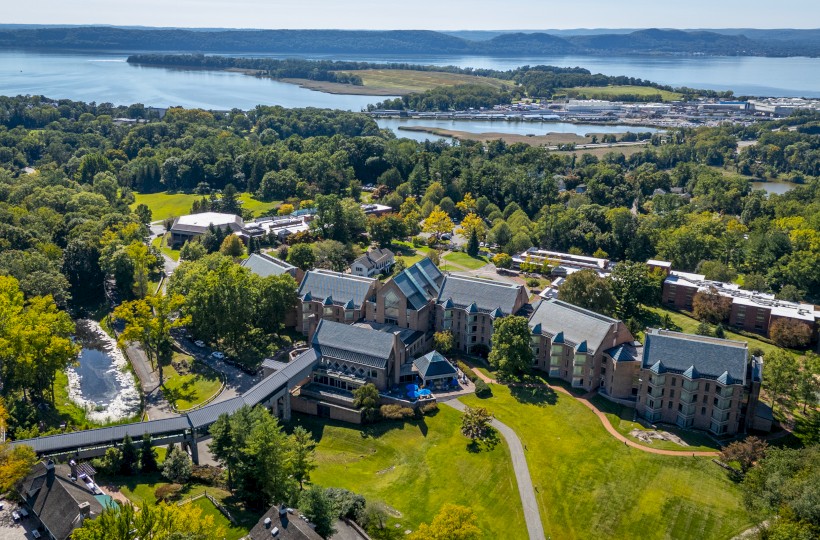 Aerial view of a large building complex surrounded by trees, near a body of water with a distant shoreline and a marina.