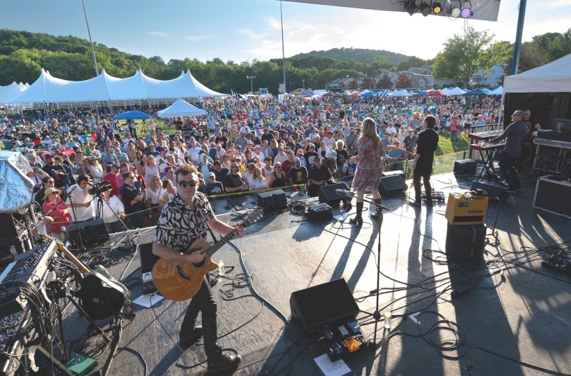 A band performs on stage at an outdoor music festival, with a large audience and tents in the background under a clear blue sky.