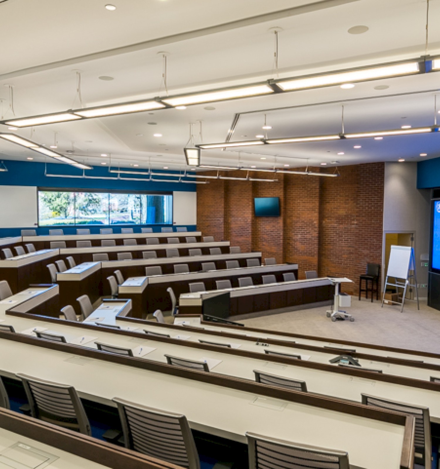 This is an empty modern lecture or conference room with tiered seating, a large digital screen, and a brick accent wall.