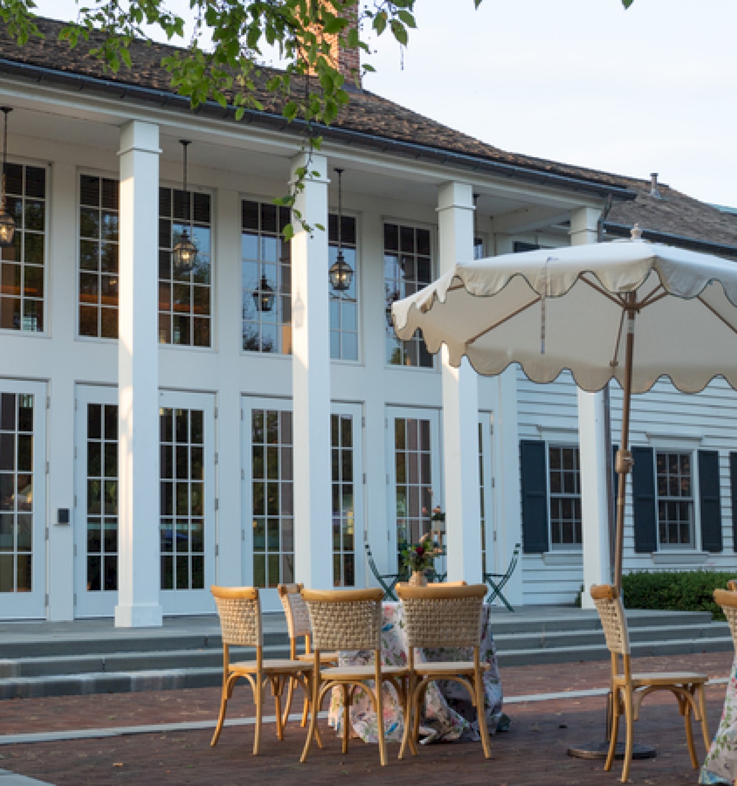 Outdoor cafe set in front of a white colonial-style building with tall doors, glass windows, and a large umbrella over wicker tables and chairs.