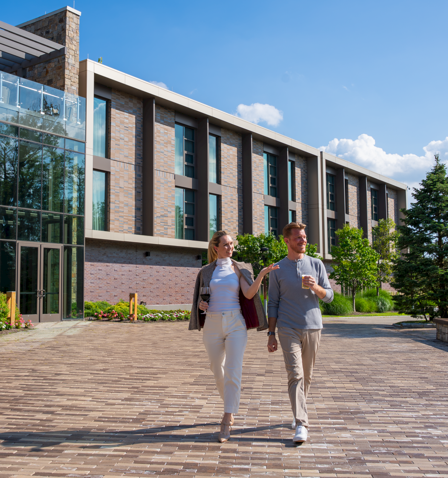 A man and woman walk and chat outside a modern building on a sunny day, holding drinks and enjoying each other's company.
