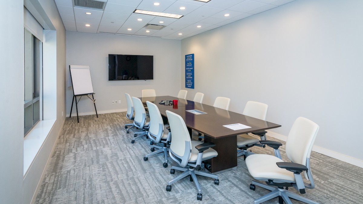 A modern conference room with a long table, eight chairs, a TV screen, a flip chart, and ample lighting.