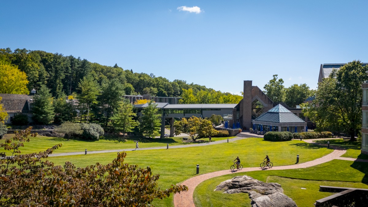 A peaceful outdoor scene with cyclists on a winding brick path, surrounded by trees, green lawns, and buildings under a clear blue sky.