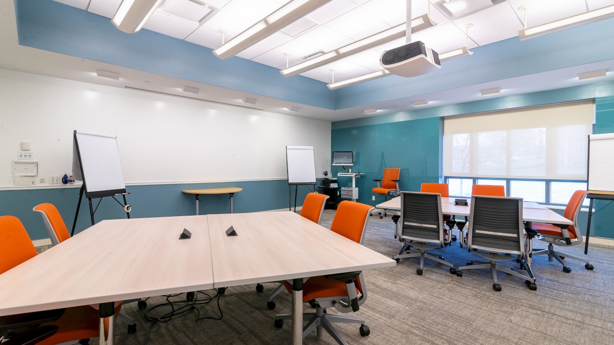 A modern conference room with tables, orange and black chairs, whiteboards, flip charts, a projector, and a large window is shown.