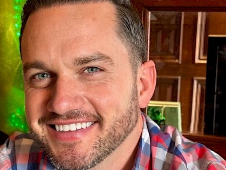 A man smiling, wearing a plaid shirt, with a wooden interior background.