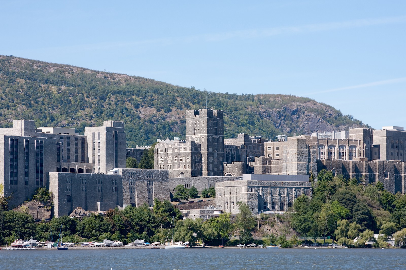 A large stone building complex is set against a hilly backdrop by a body of water, surrounded by greenery and under a clear sky.