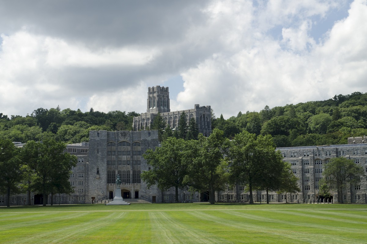 A large stone building surrounded by trees, with a green lawn in the foreground, situated under a partly cloudy sky.