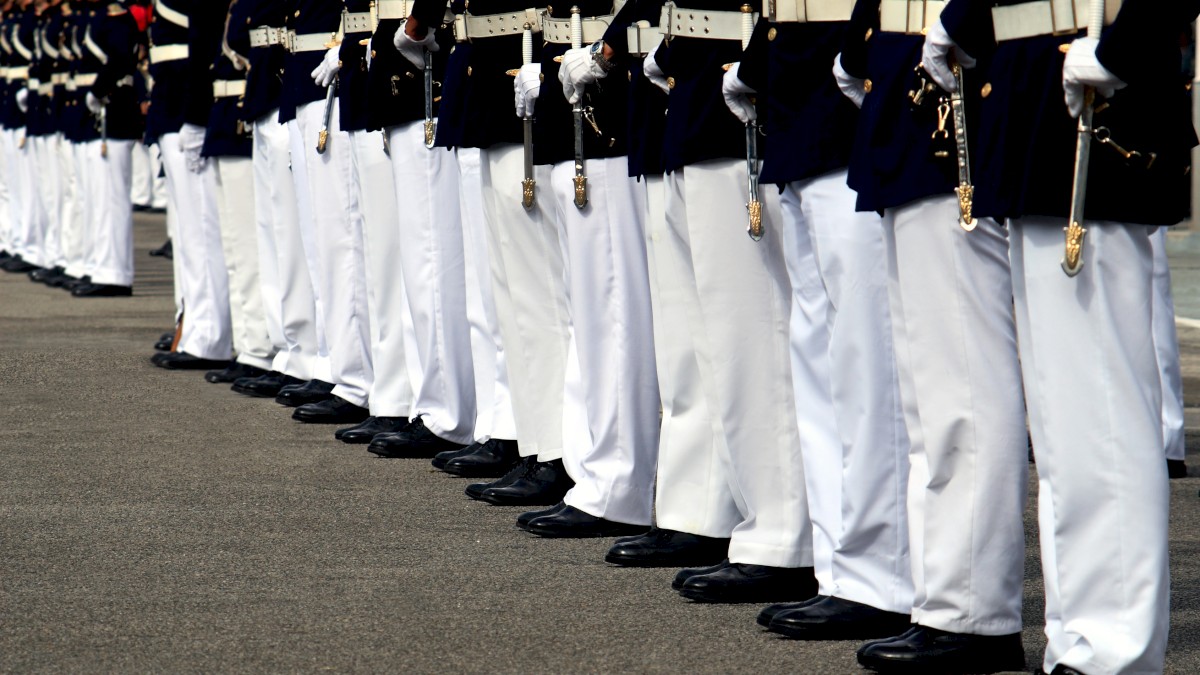 A row of military personnel in uniform stands with precision, wearing dark jackets, white pants, black shoes, and holding swords.
