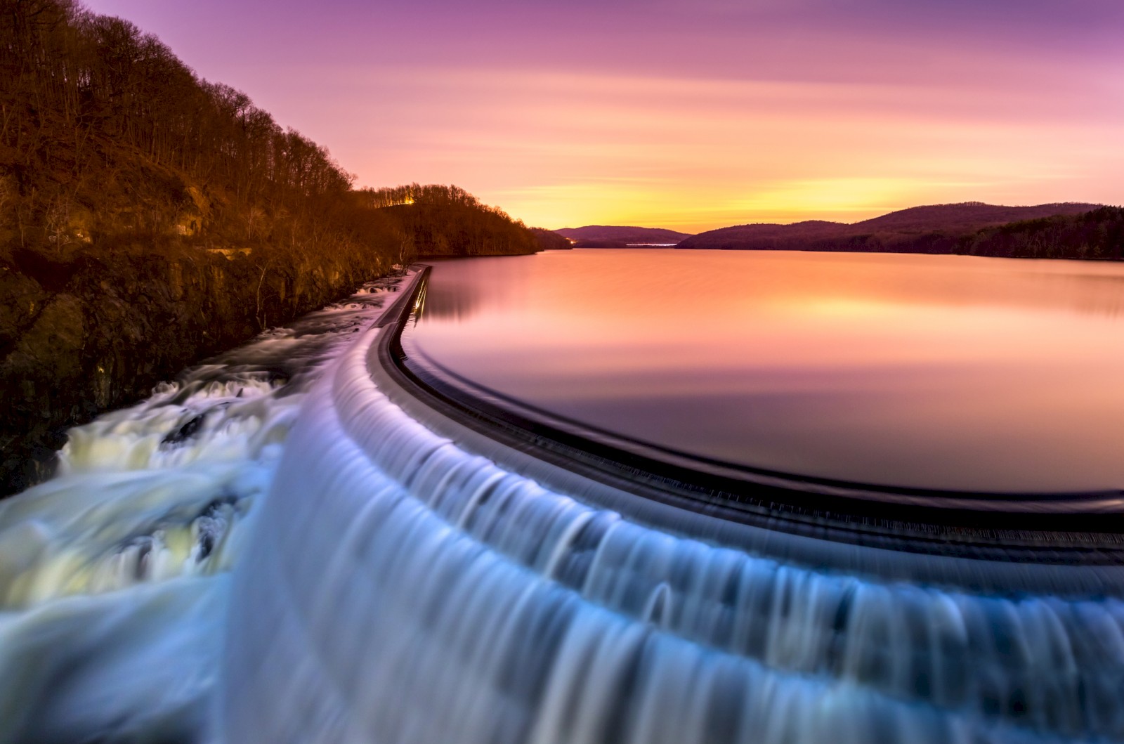 A stunning sunset over a serene reservoir with a smooth waterfall, surrounded by trees and hills, creating a peaceful landscape.