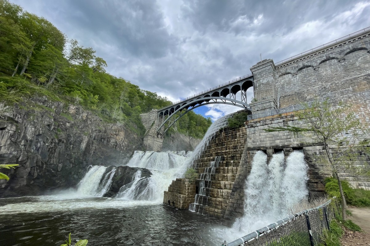 A cascading waterfall flows beneath an arched bridge, surrounded by lush greenery, under a partly cloudy sky.
