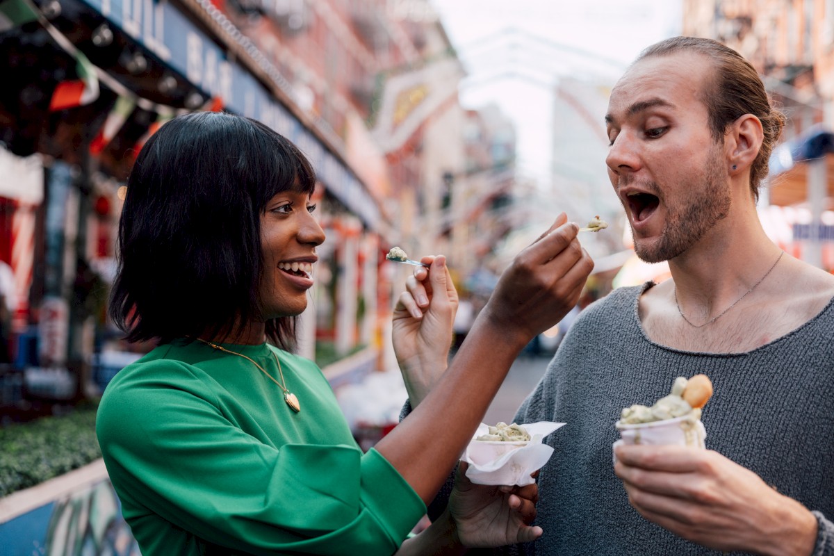A woman and man are joyfully feeding each other ice cream on a city street, with colorful buildings in the background.