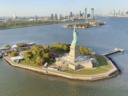 The image shows the Statue of Liberty on Liberty Island with water and a distant city skyline in the background.