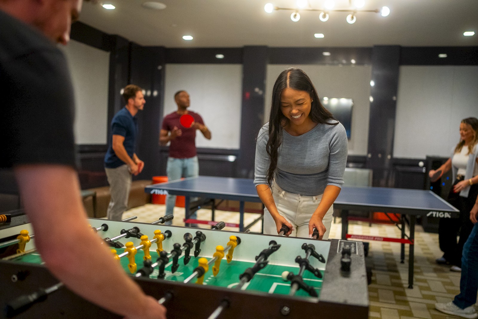 People are enjoying a game room, playing foosball and table tennis.
