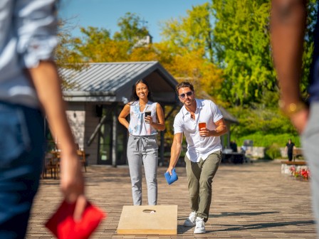People are playing cornhole outdoors, with one person tossing a bean bag towards the board. Others look on in a relaxed setting.