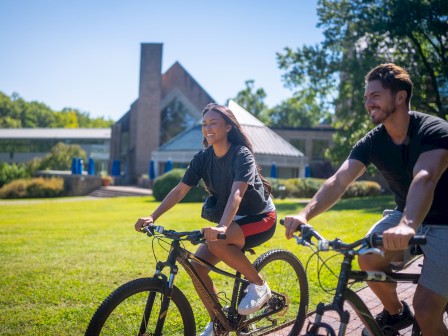 Two people are riding bicycles on a grassy area with trees and a large building in the background.