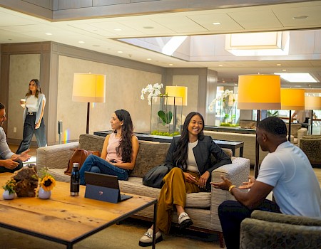 A group of people are sitting in a modern living room, chatting and enjoying drinks, with one person walking by in the background.