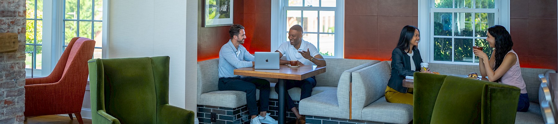 People in a cozy, modern room with wood accents are chatting around tables, with some using laptops and others engaged in conversation.