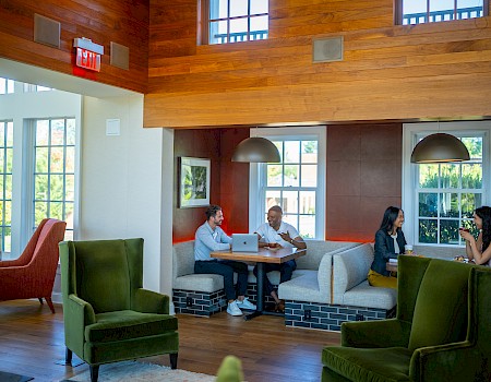 People in a cozy, modern room with wood accents are chatting around tables, with some using laptops and others engaged in conversation.