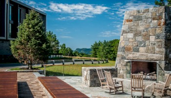 The image shows an outdoor area with a large stone fireplace, wooden chairs, a table, trees, a building, and a scenic background under a blue sky.