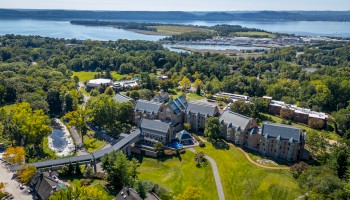 Aerial view of a large building complex surrounded by trees, near a body of water with a distant shoreline and a marina.