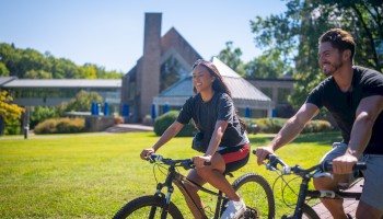 Two people are riding bicycles on a grassy path with a modern building in the background and trees around.