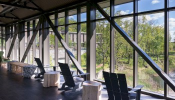 A spacious hallway with large windows shows a scenic outdoor view, including trees and a pond with fountains, featuring seating arrangements.