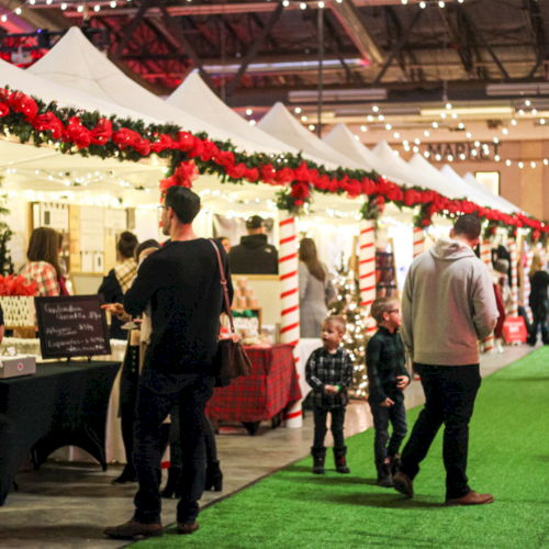 A festive indoor market scene with decorated stalls and people browsing; the atmosphere is lively with string lights overhead.