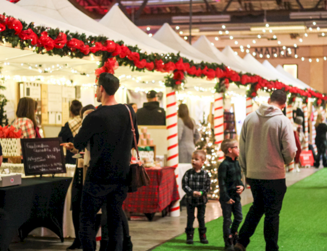 A festive indoor market scene with decorated stalls and people browsing; the atmosphere is lively with string lights overhead.