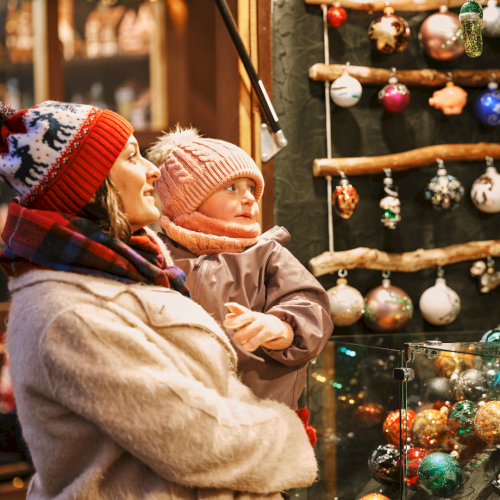 A woman and child in warm clothing admire Christmas ornaments displayed at a festive market stall, filled with colorful decorations.