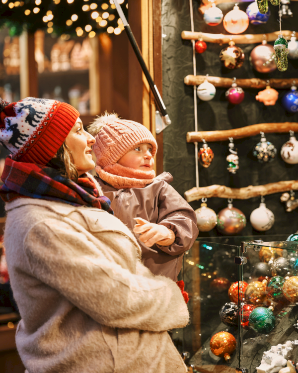 A woman and child in warm clothing admire Christmas ornaments displayed at a festive market stall, filled with colorful decorations.