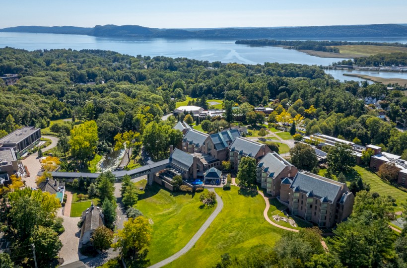 Aerial view of a lush landscape with a large building complex surrounded by trees, overlooking a body of water and distant hills.