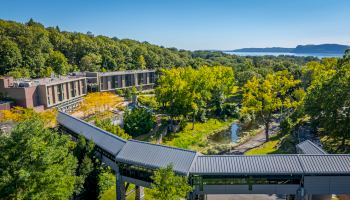 A scenic view of a modern building surrounded by lush greenery and a river, with hills and a waterbody in the background.