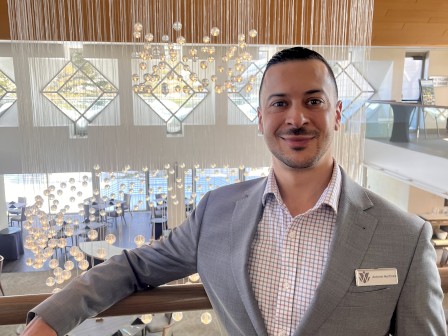 A man in a light gray suit stands in a stylish, modern indoor space with a large circular chandelier above.