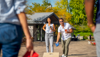 People playing cornhole outside, with a man tossing a bean bag toward the board. Others are watching, holding red bags in hand.