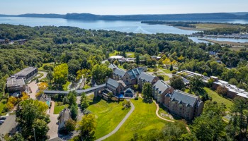 Aerial view of a lush landscape with a large building complex surrounded by trees, overlooking a body of water and distant hills.