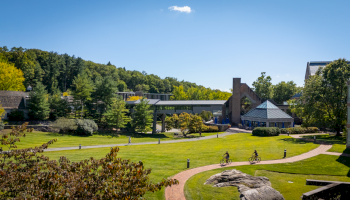 A scenic campus view features pathways, green lawns, trees, and people biking under a clear blue sky.