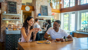 A couple enjoys coffee and pastries in a cozy café with brick walls and large windows, creating a warm and inviting atmosphere.