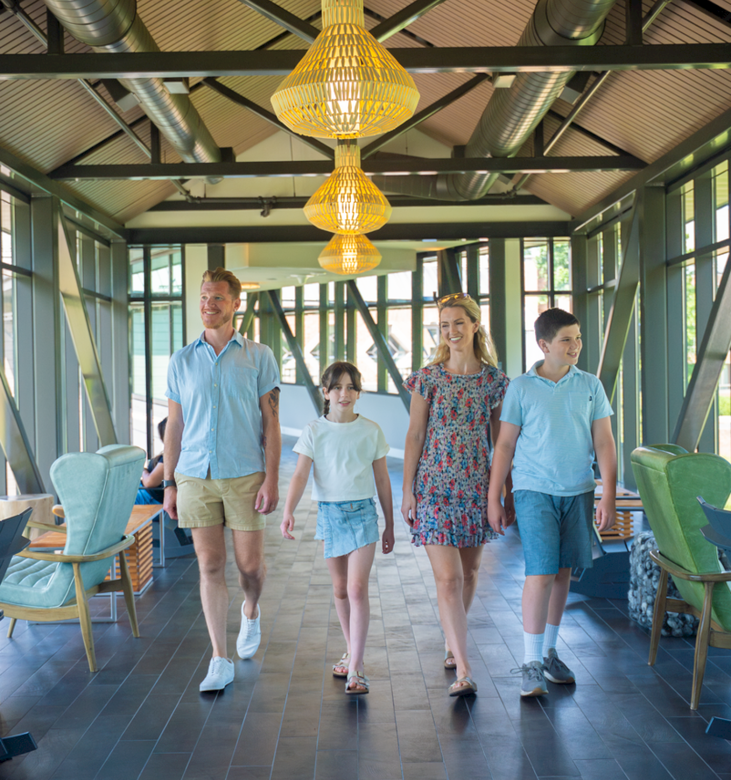 A family of four walks happily inside a modern, brightly-lit cafe or restaurant with large windows and stylish yellow light fixtures.
