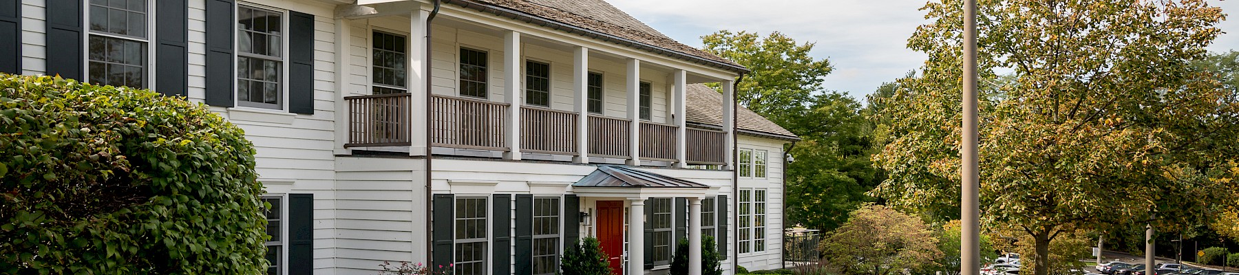 A large white house with black shutters, a brick walkway, landscaped bushes, and a street lamp in a lush, green setting.