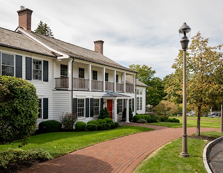 A large white house with black shutters, a brick walkway, landscaped bushes, and a street lamp in a lush, green setting.