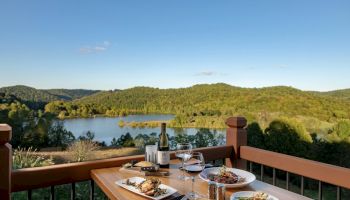 A wooden table with food, wine, and glasses set for two, overlooking a scenic lake and green hills under a clear blue sky, is pictured.