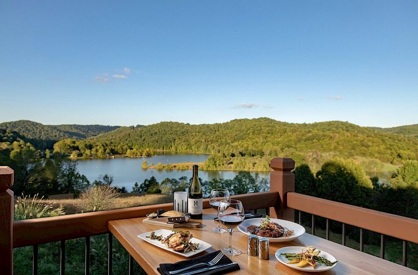 A wooden table with food, wine, and glasses set for two, overlooking a scenic lake and green hills under a clear blue sky, is pictured.