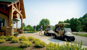 People preparing for a golf round, with two golf carts, lush greenery, and a wooden building with a green roof in the background.