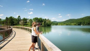 Two people stand on a wooden boardwalk overlooking a calm lake surrounded by lush green trees and hills under a clear blue sky.